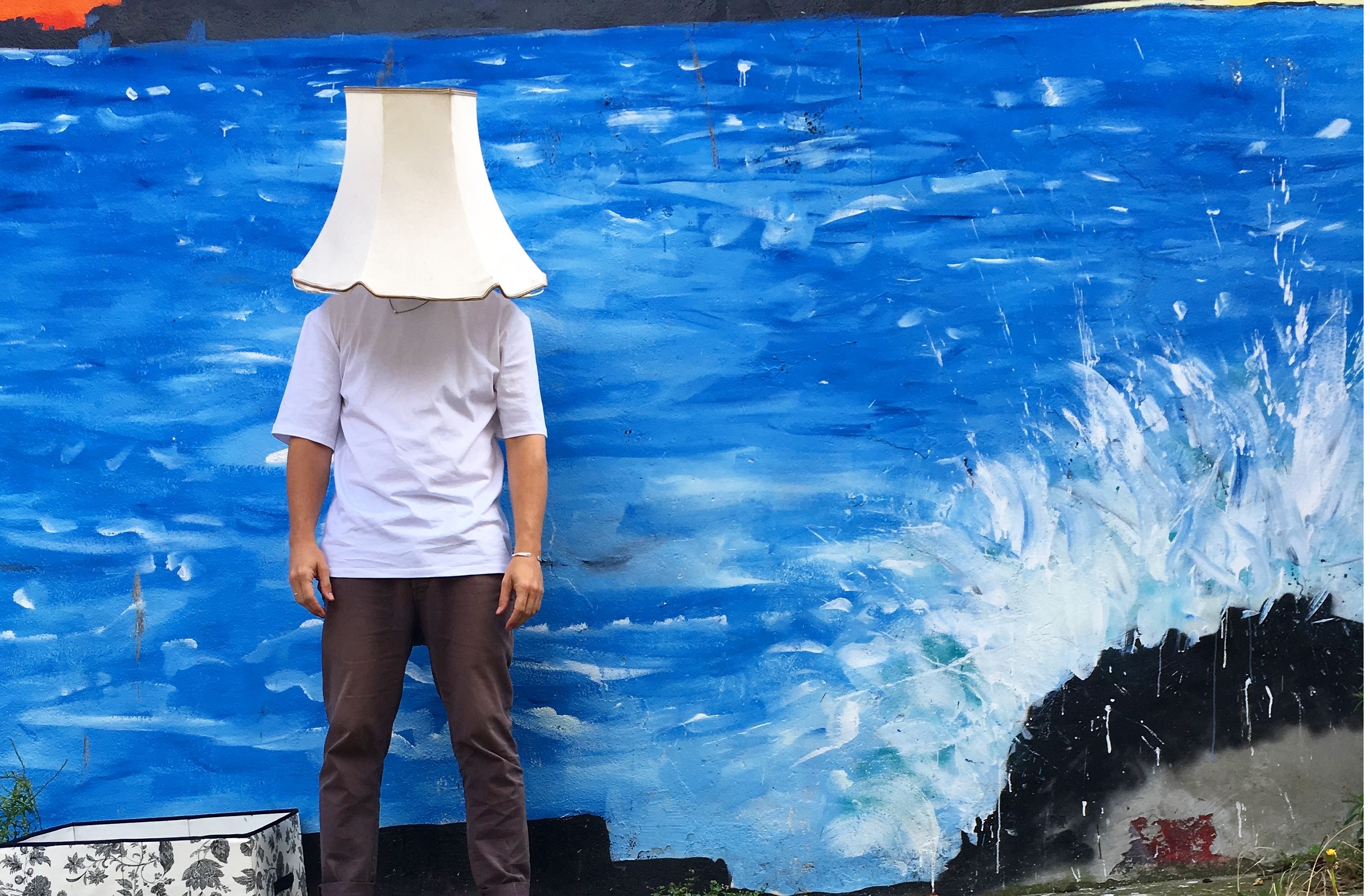 The artist wears a white retro lampshade on his head, in front of a vibrant blue-painted wall.