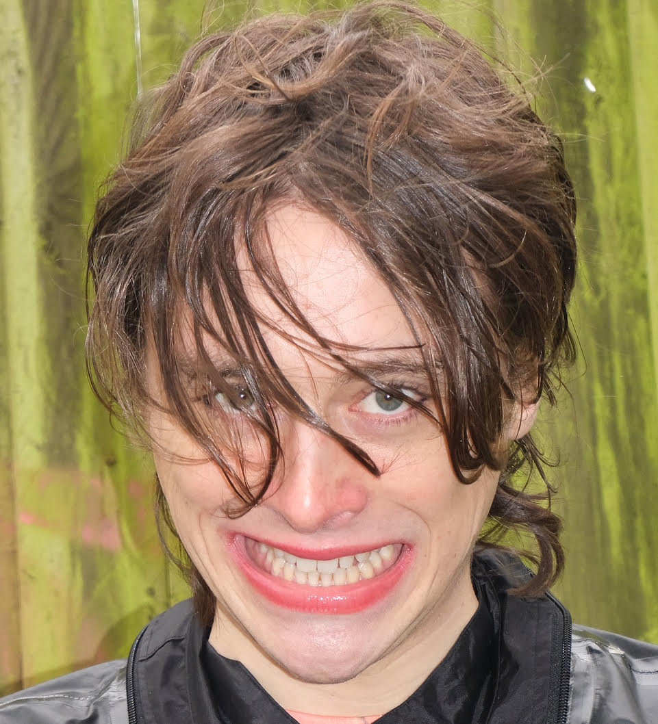 The artist bares teeth at the camera, against an industrial green background.